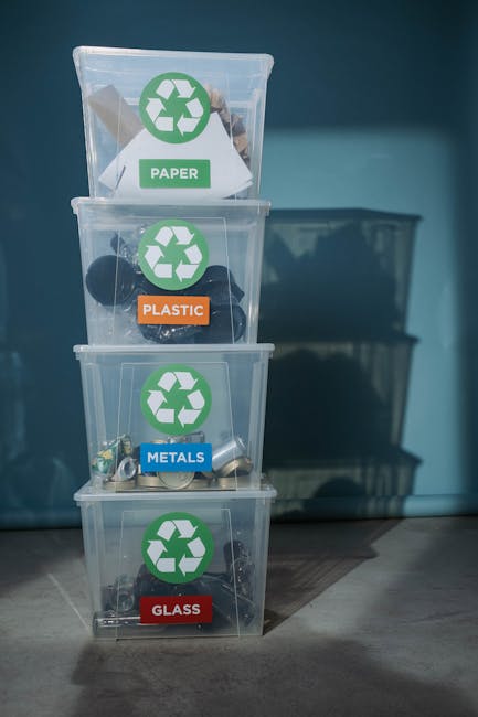A set of four transparent plastic recycling bins stacked vertically against a light blue wall, each labeled with a coloured sticker and recycling symbol to indicate the type of waste: the top bin with