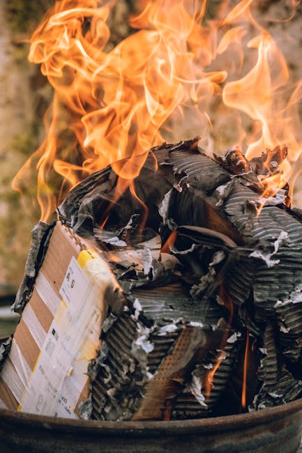 A close-up of a metal drum filled with tightly packed cardboard packaging, some of which is charred and blackened, with flames rising and flickering above the materials. The cardboard appears to be co